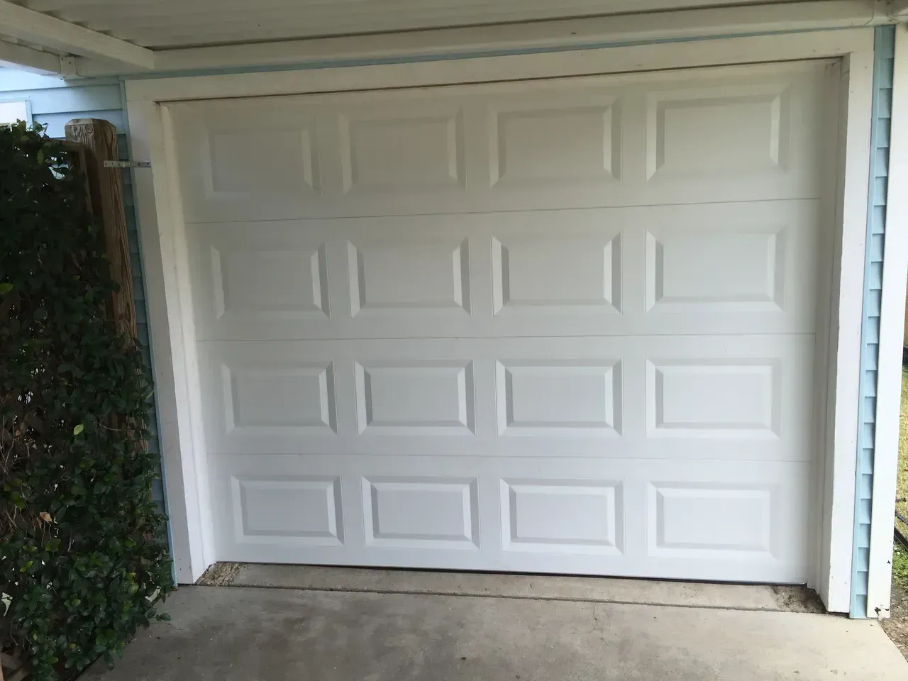 White garage door on a concrete driveway, next to blue siding and bushes.