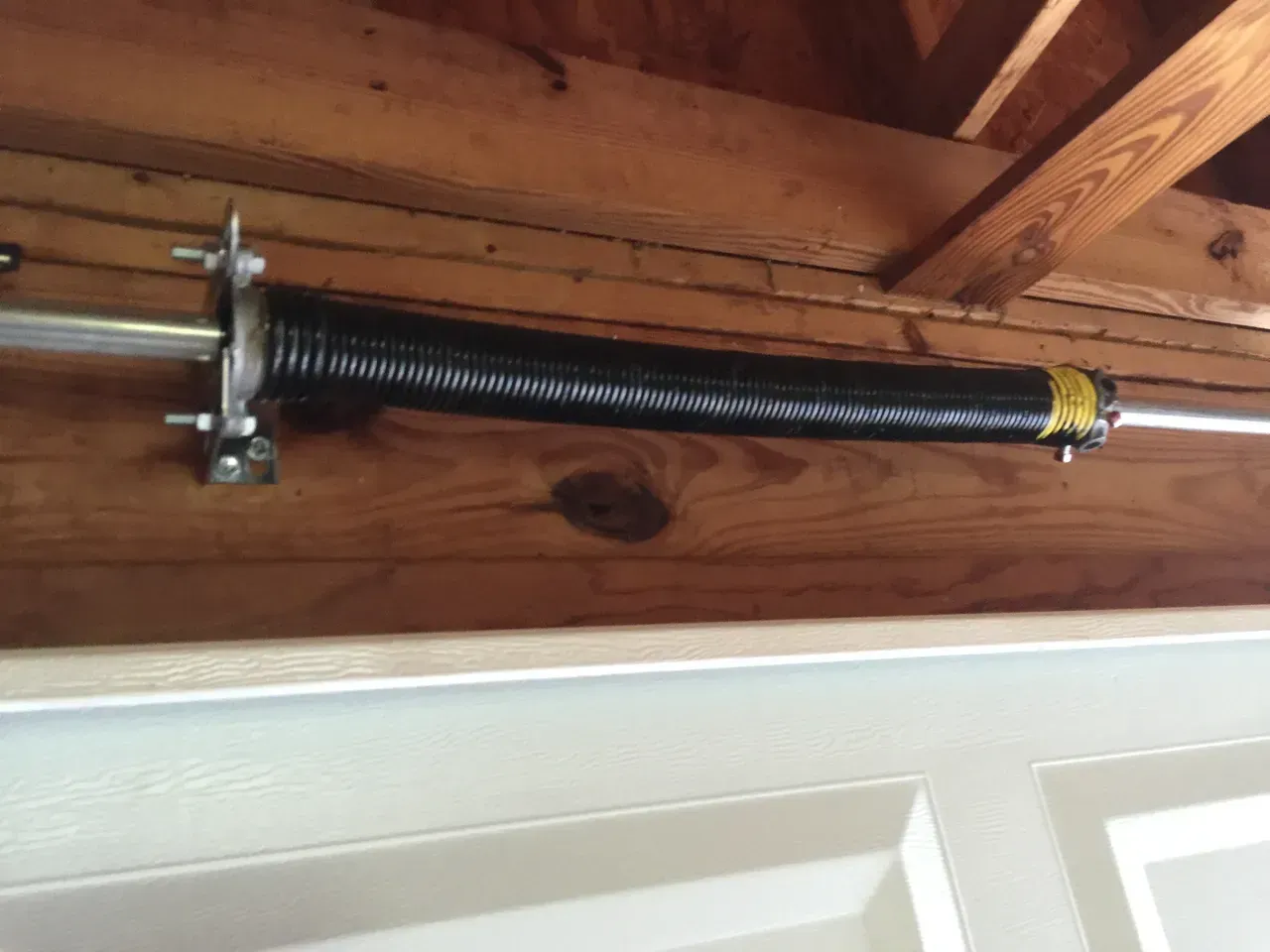 Garage door torsion spring, black coiled, mounted above white garage door. Wooden beams in the background.