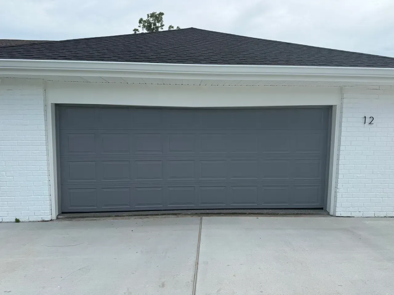 Gray garage door on white brick building, number 12 visible. Concrete driveway below, overcast sky.