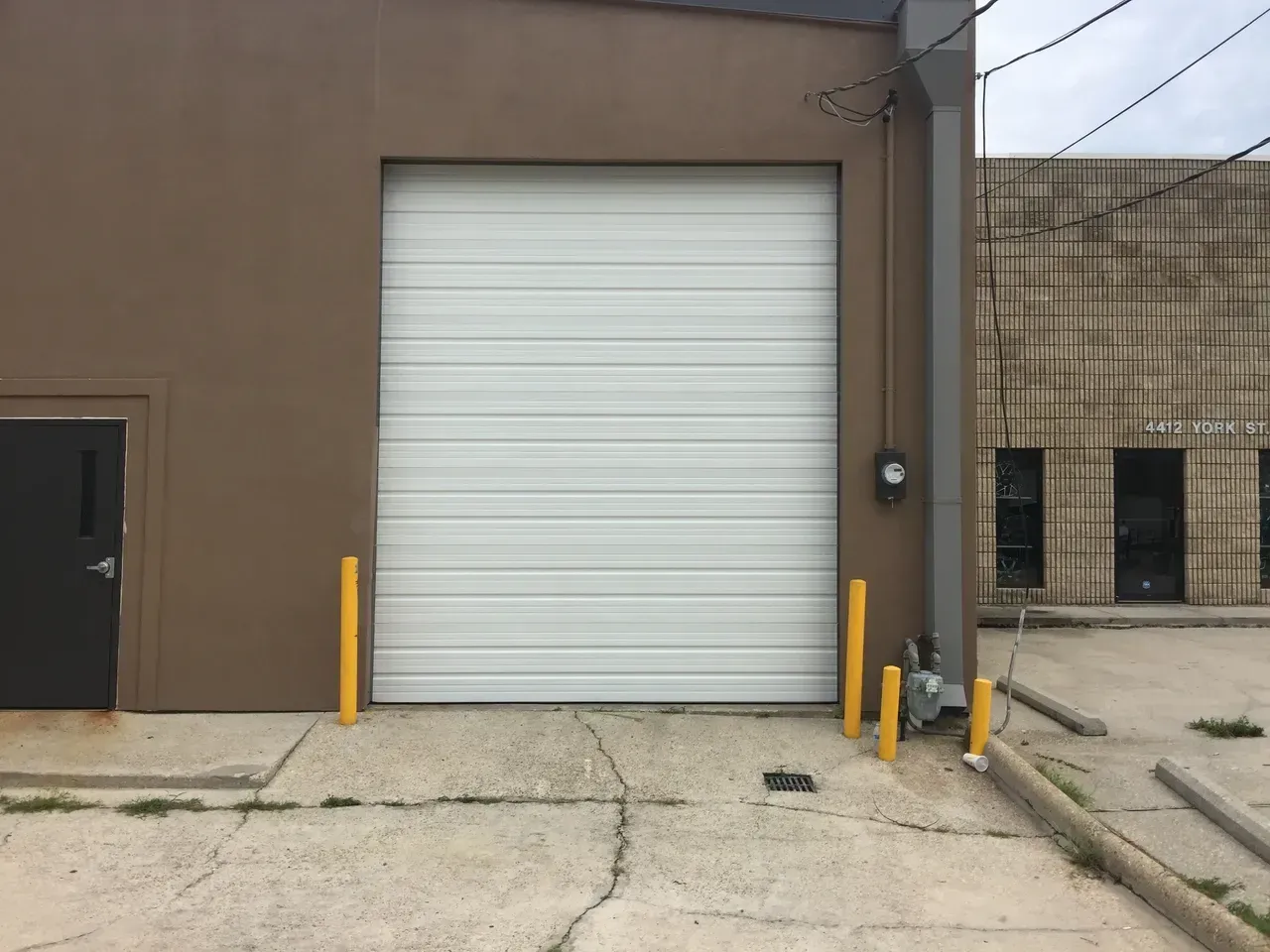 Brown building with closed white garage door, yellow bollards, concrete ground.
