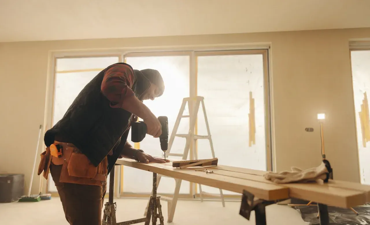Person using a drill on a wooden board at a work table in a room with large windows; ladder in background.