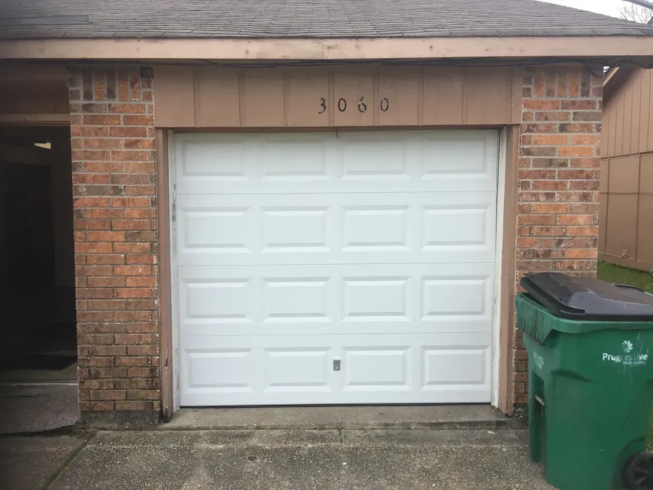 White garage door, brick building with green trash can and address number 3036.
