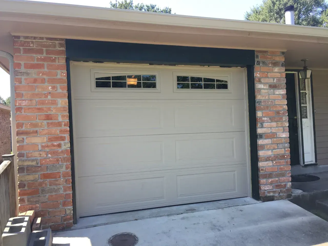 Tan garage door with small windows, brick exterior, and dark trim.