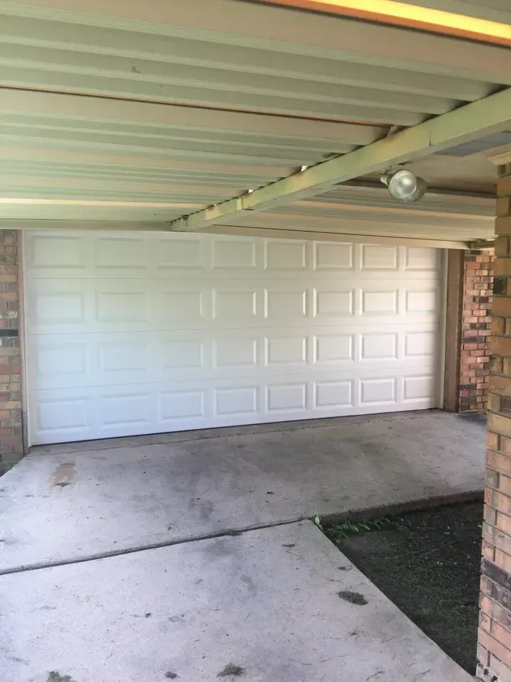 White garage door under a carport, set on a concrete driveway. Brick pillars support the roof.