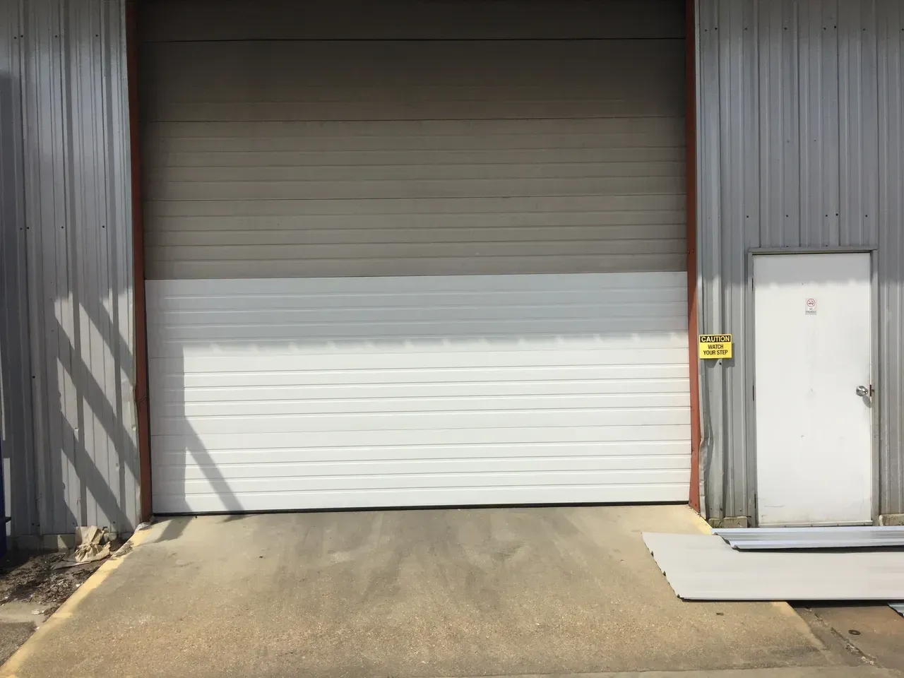 White closed garage door with a brown frame, gray building exterior, and a small white door on the right.