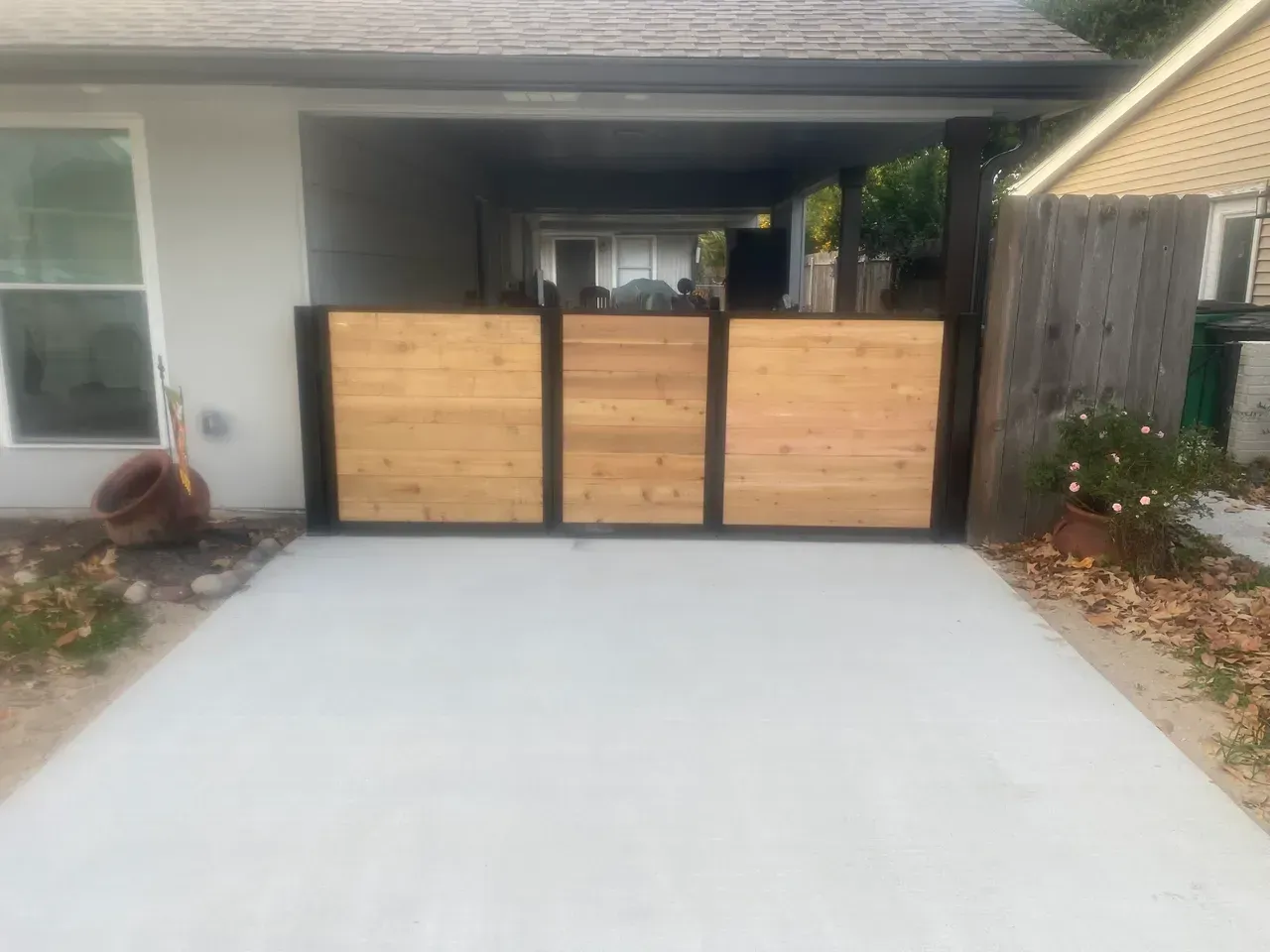Concrete driveway leads to a covered area with a wood and metal gate.  Two potted plants are visible.