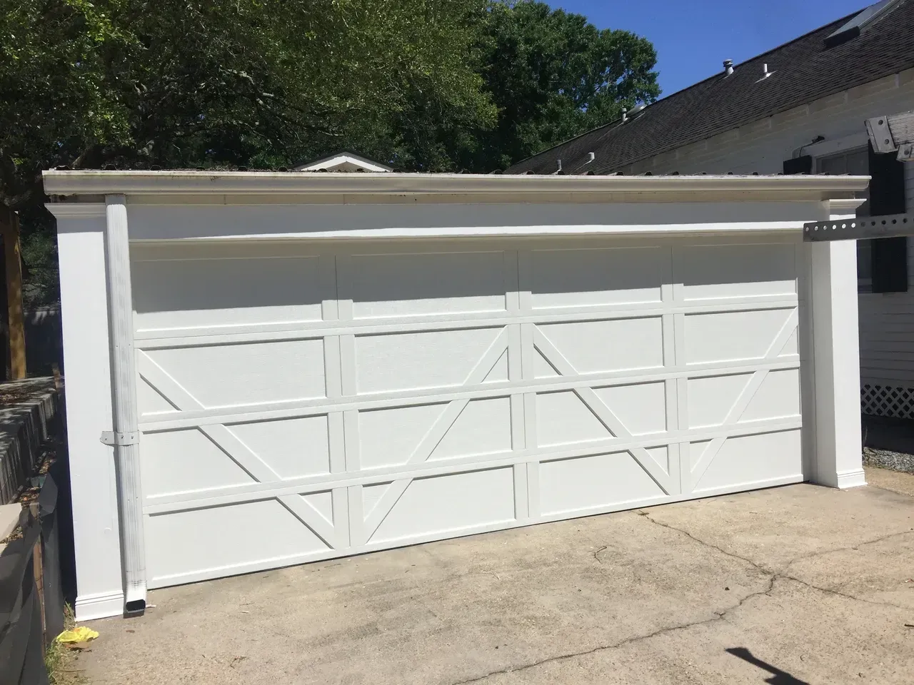 White garage door with an X-pattern design. Concrete driveway, white building trim, and greenery in the background.