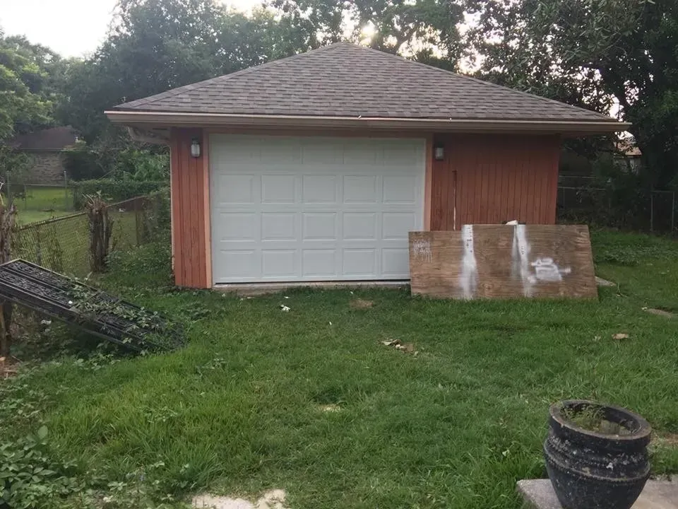 Small tan garage with white door, on a grassy lot. A board rests in front. Trees in the background.