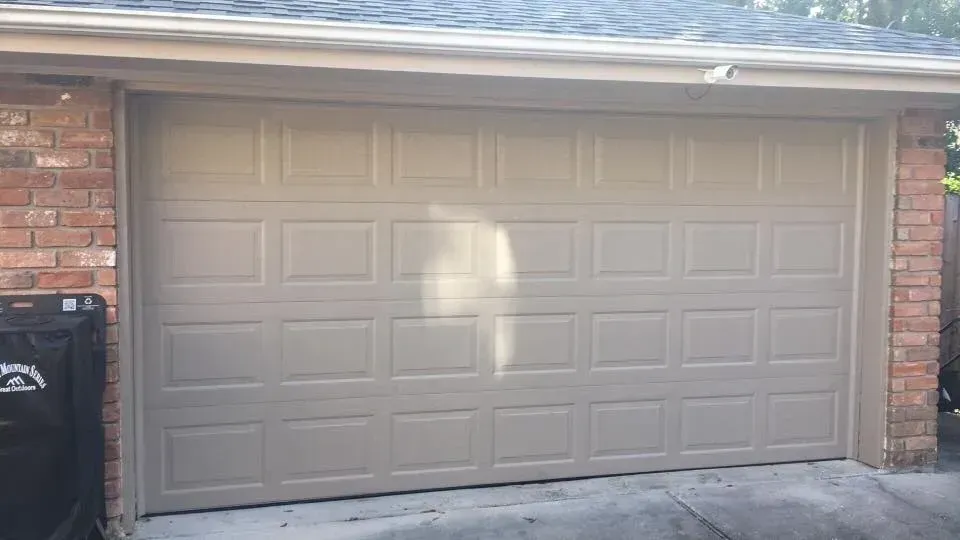 Beige panel garage door on a brick building with a black trash can on the left.