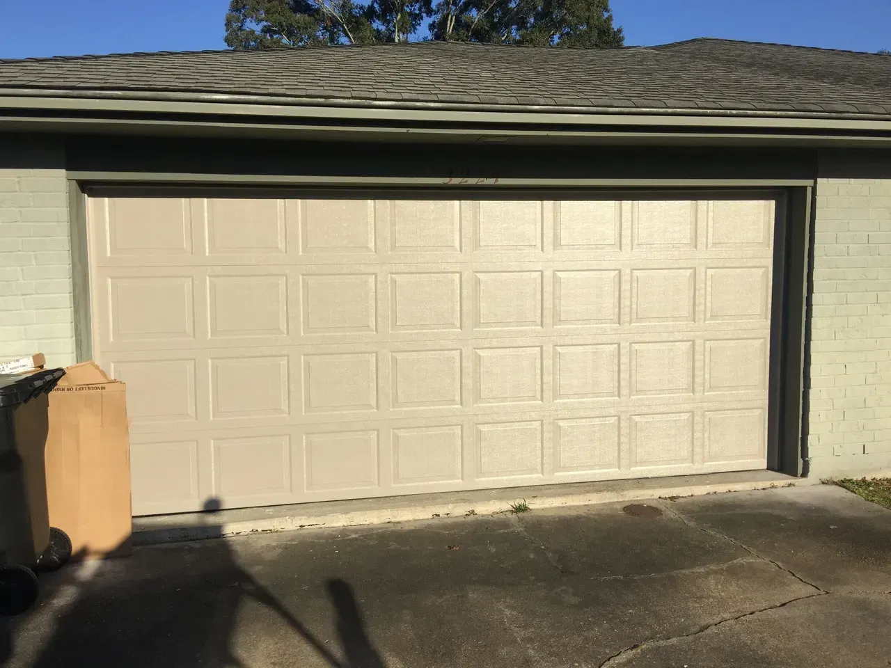 Beige panel garage door on a light green brick building; cardboard box on left.