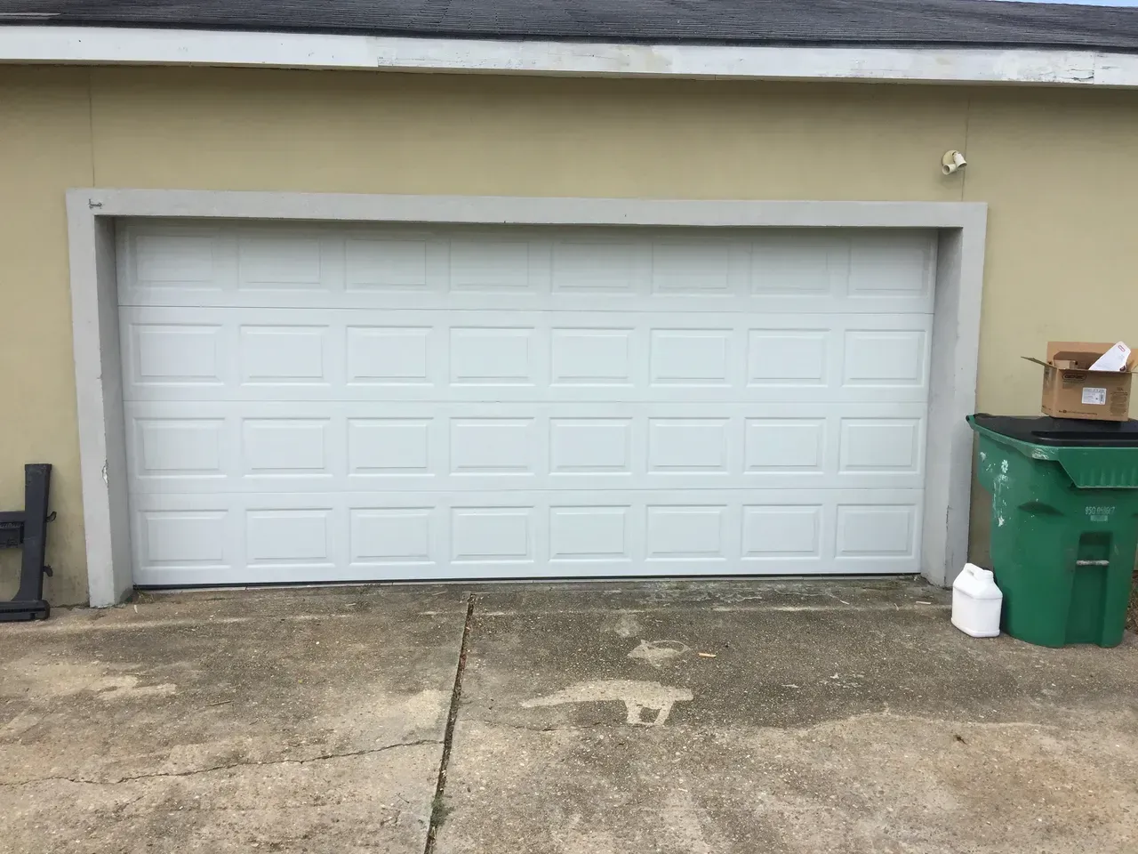 White garage door on a beige building with a cracked concrete driveway. A green trash bin and cardboard box are nearby.