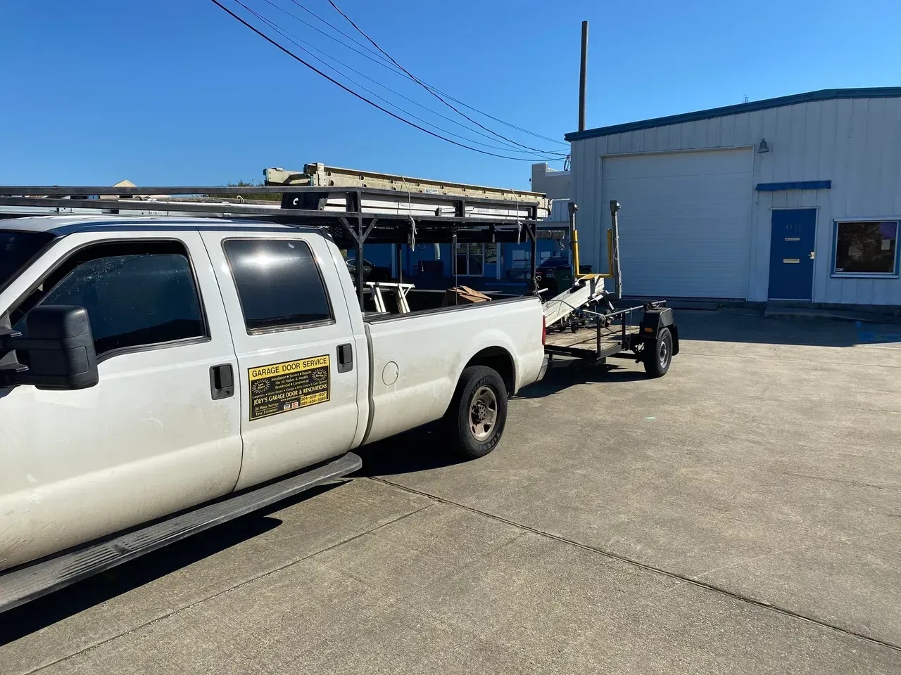 White pickup truck with a trailer parked in front of a blue-doored building on a sunny day.