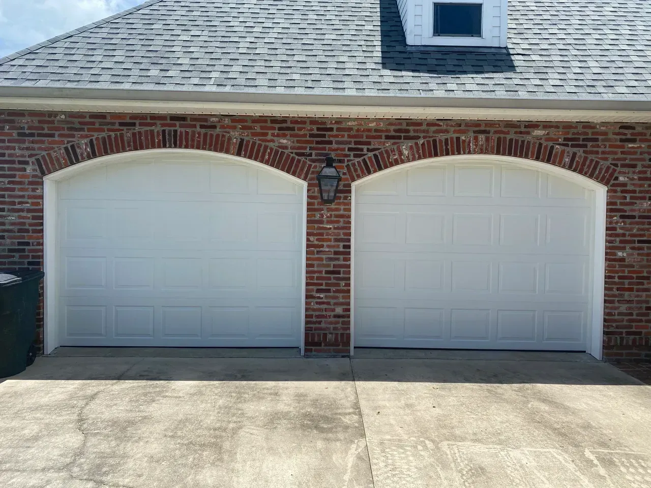 Two white garage doors with arched brick detail, brick building, asphalt driveway, gray roof.