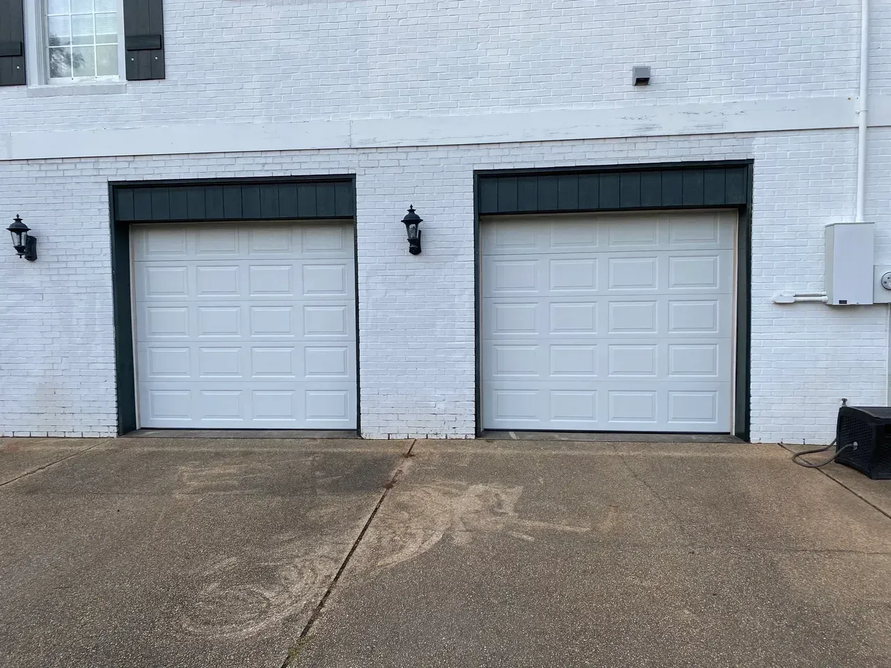 Two white garage doors with black trim, mounted on a white brick building.