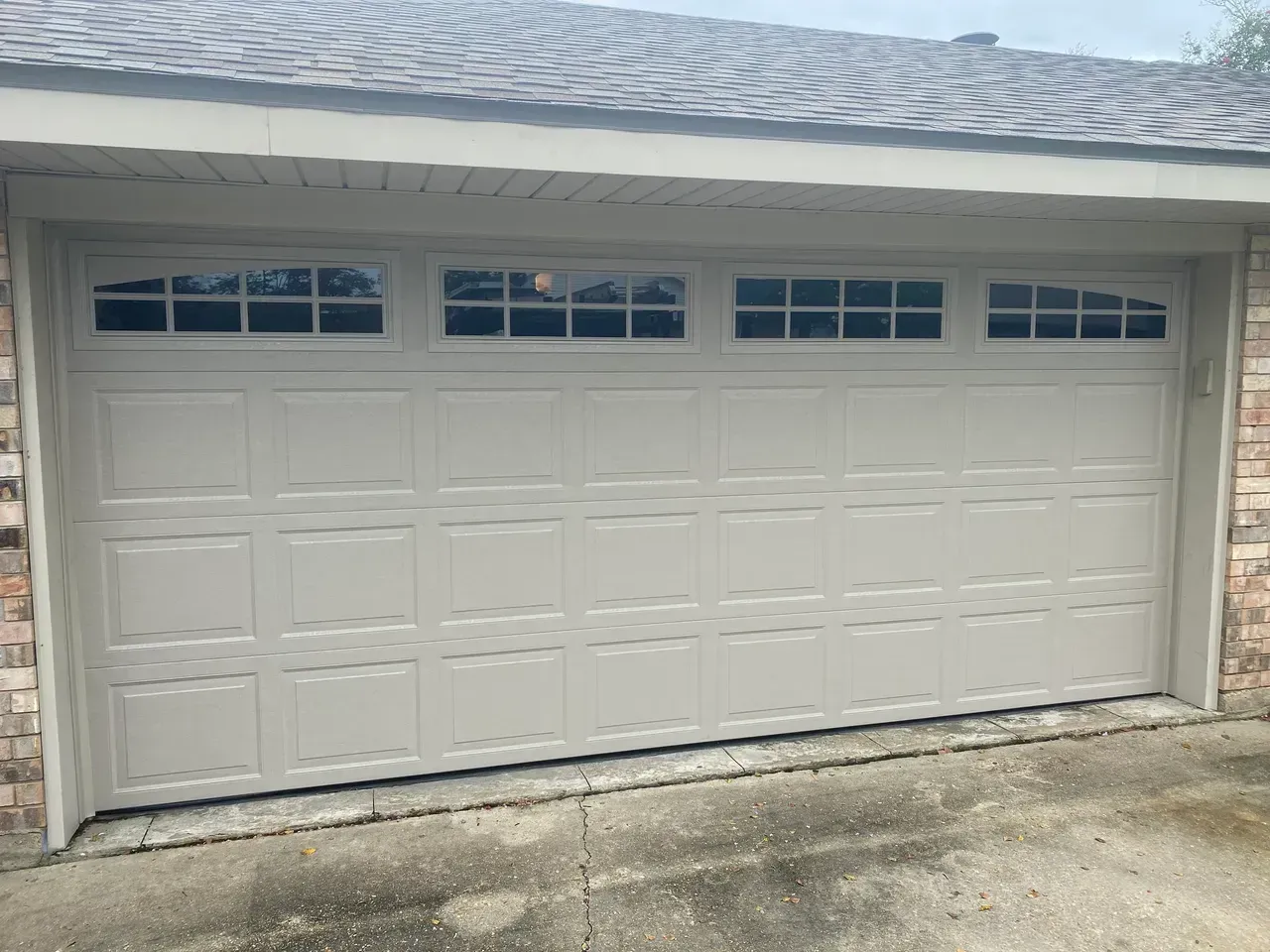 Tan garage door with small rectangular windows at the top. Brick siding and overcast sky.