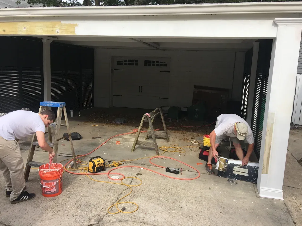 Two people renovating a garage, one mixing paint, the other working inside a toolbox; tools and materials are visible.