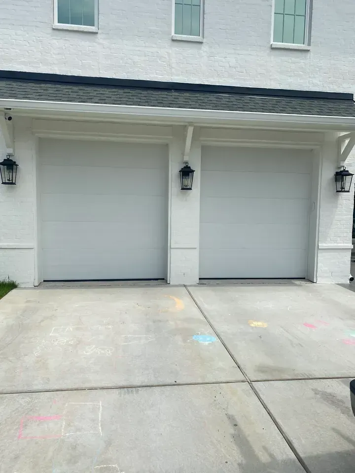 Two white garage doors on a brick house with exterior lights. Concrete driveway has colorful chalk marks.
