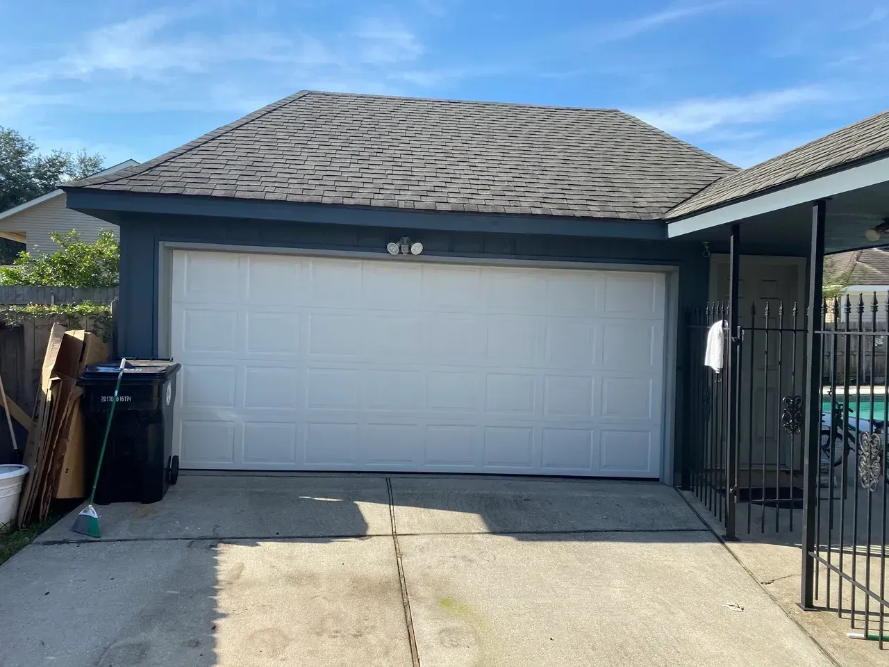 Garage with white door, gray roof, and blue siding. Black trash can to the left and black gate to the right.