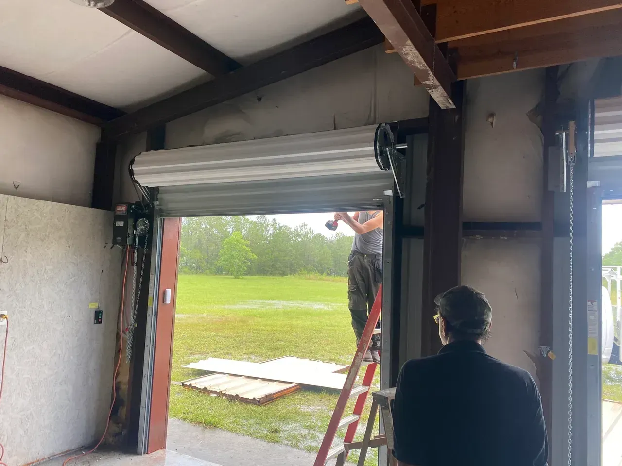 Man on ladder installing a metal roll-up garage door in a building with a view of a rainy field.