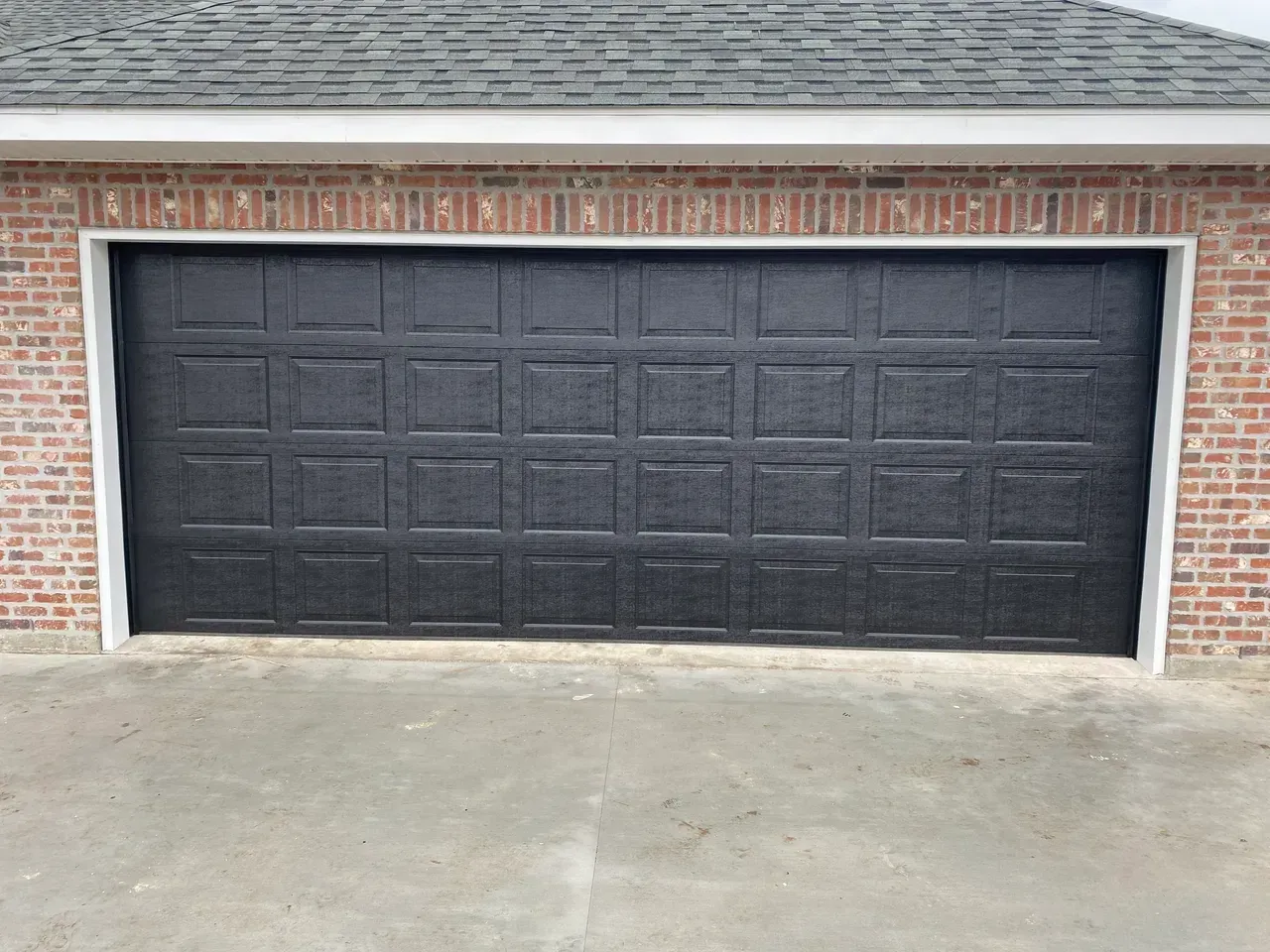 Black garage door with square panels, set in brick building with concrete driveway.