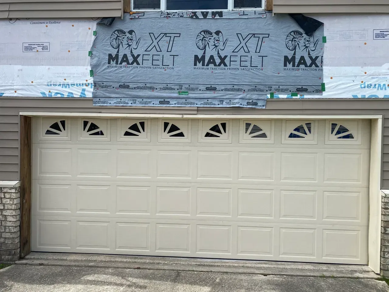Beige garage door with arched glass panels, beneath siding and house wrap.