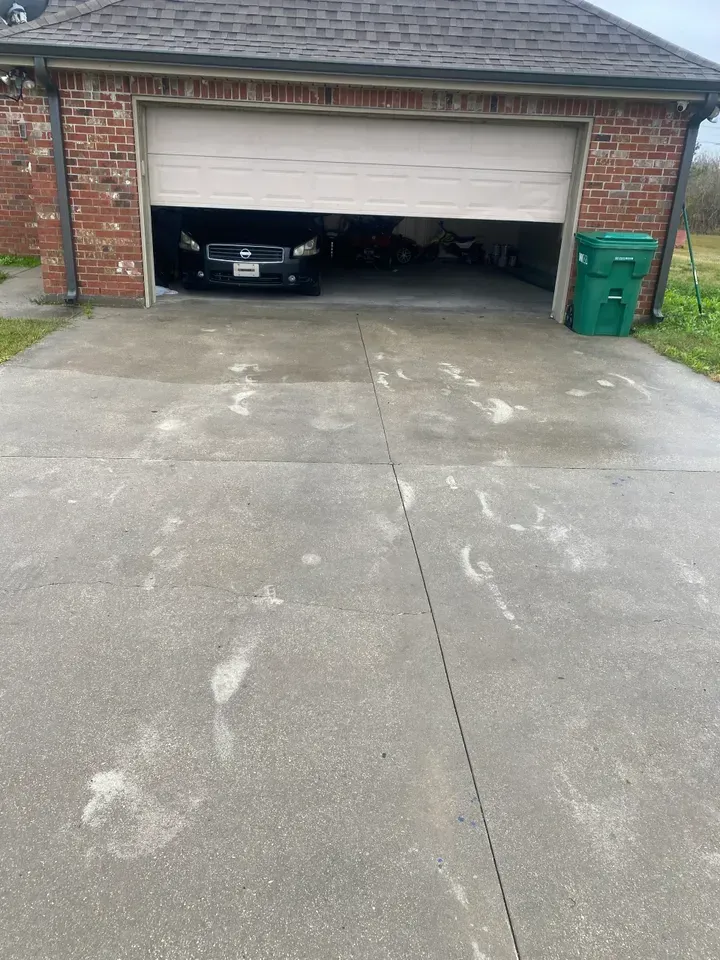 Garage with car parked inside, partially open door, concrete driveway. Green trash can on right.