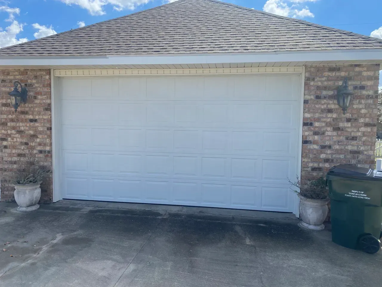 White garage door on a brick building with decorative sconces, potted plants, and a green trash can.