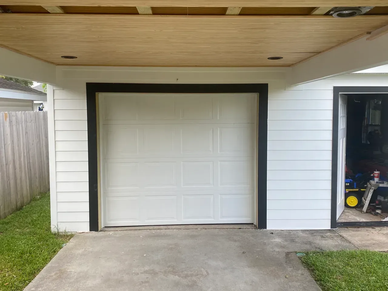 White garage door framed in black, under a wooden ceiling. Concrete driveway, green grass, and a door to the right.