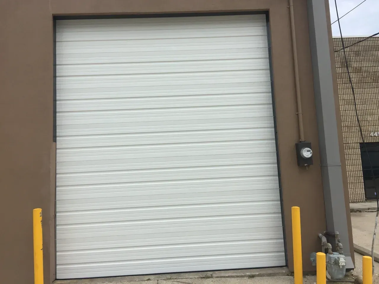 White closed garage door in a brown building, flanked by yellow bollards, electrical box visible.