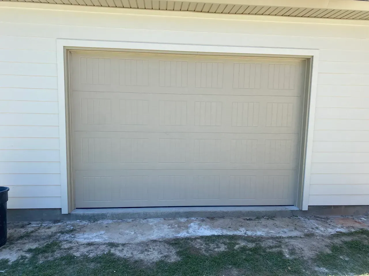 Tan garage door on a beige house, front view. Ground has grass and white residue.