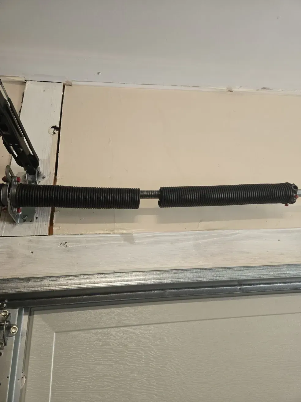 Garage door torsion spring, black and broken, mounted above a closed door.