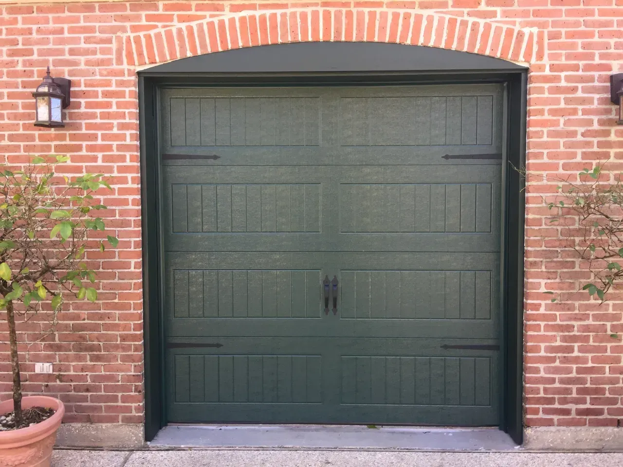 Green garage door set in a red brick wall with an arched brick top and two outdoor wall lights.