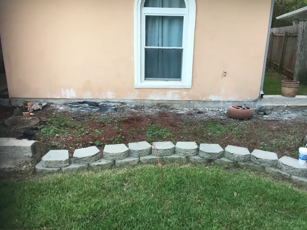 A backyard with a flowerbed bordered by gray blocks in front of a stucco building with a window.