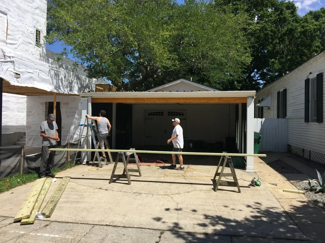 People working on construction; garage with open door; lumber on saw horses, sunny outdoors.