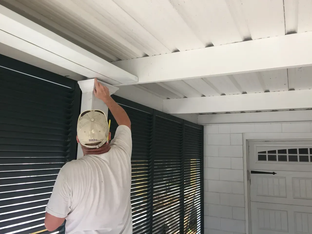 Person painting white trim on a porch, which has dark green shutters and a white ceiling.