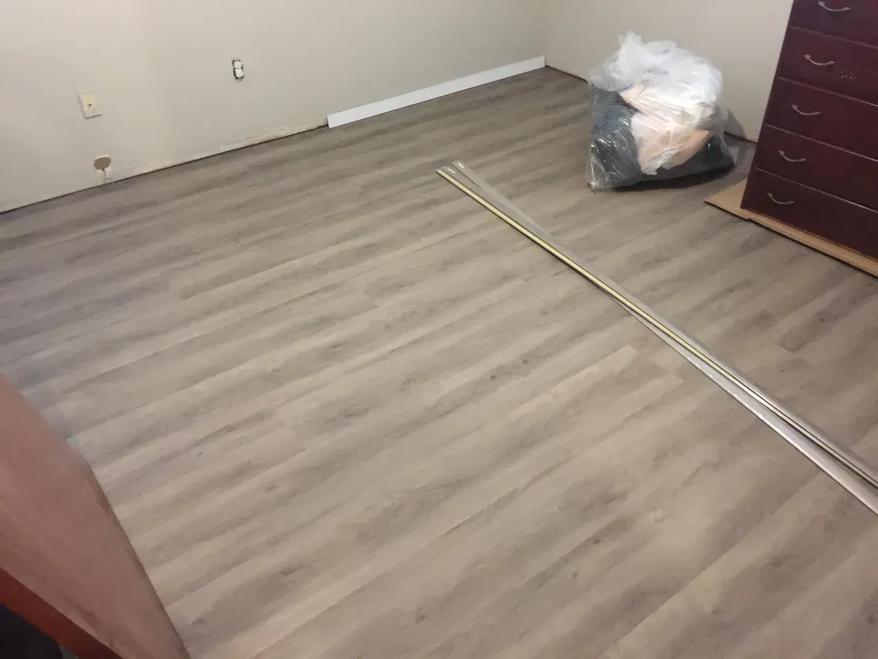 A room with light wood-look flooring and a dresser. Plastic bag and trim pieces are present.