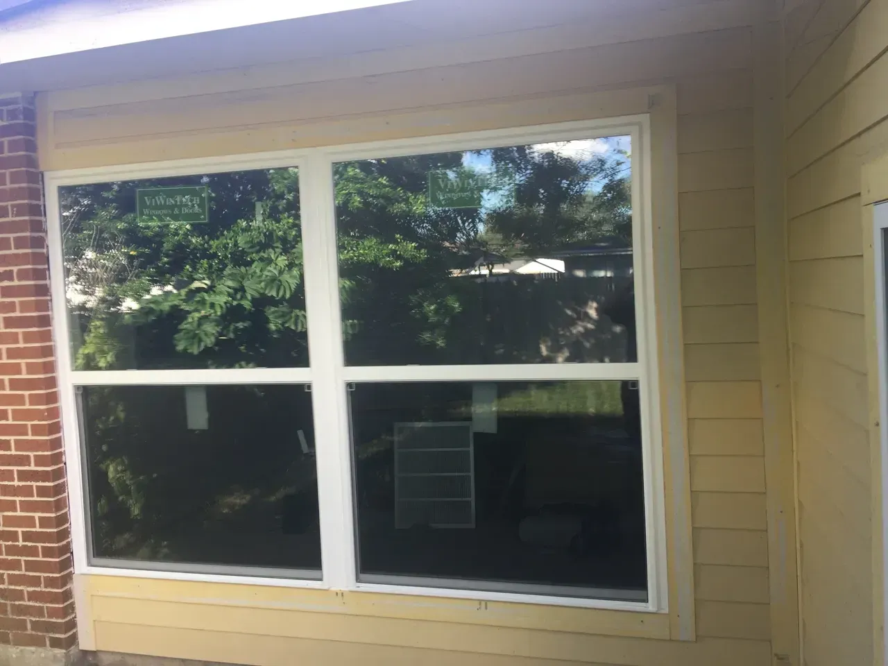 New double-hung window installed in a yellow-sided house with brick accents; the glass reflects trees.