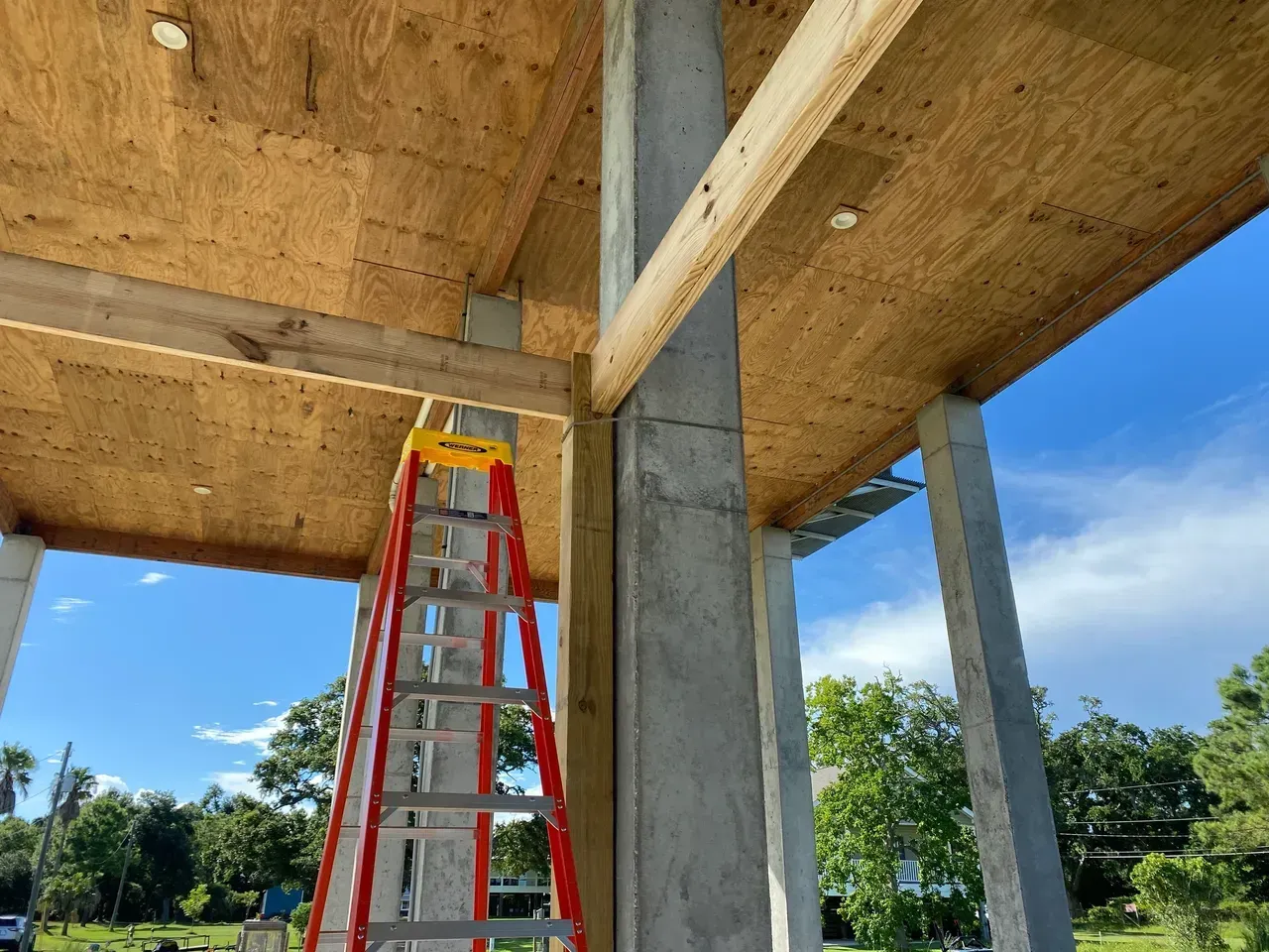 Red ladder leaning against a concrete pillar at a building under construction; wooden beams and ceiling visible.