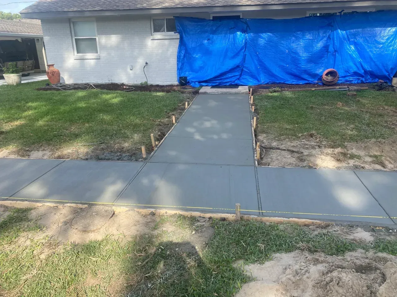 Freshly poured concrete sidewalk leading to a house with a blue tarp overhead.