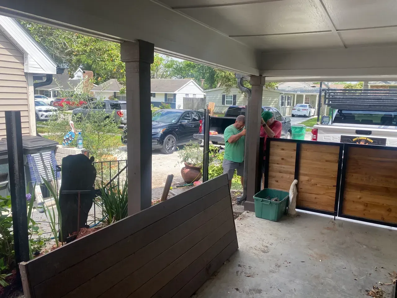 Two people installing a fence, under a porch with brown columns, a car parked in the background.