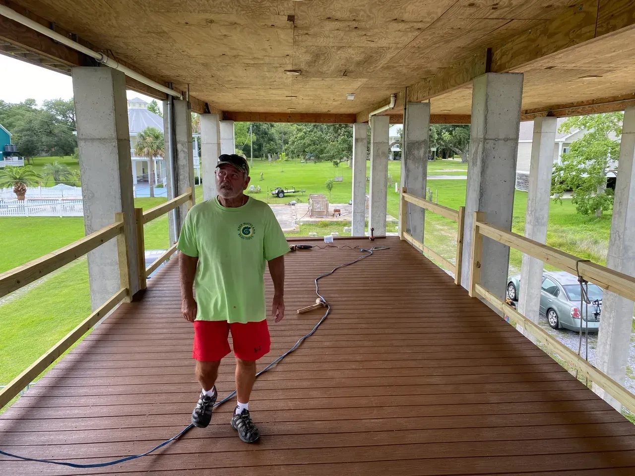 Man in red shorts and green shirt on a porch under construction, overlooking a grassy yard.
