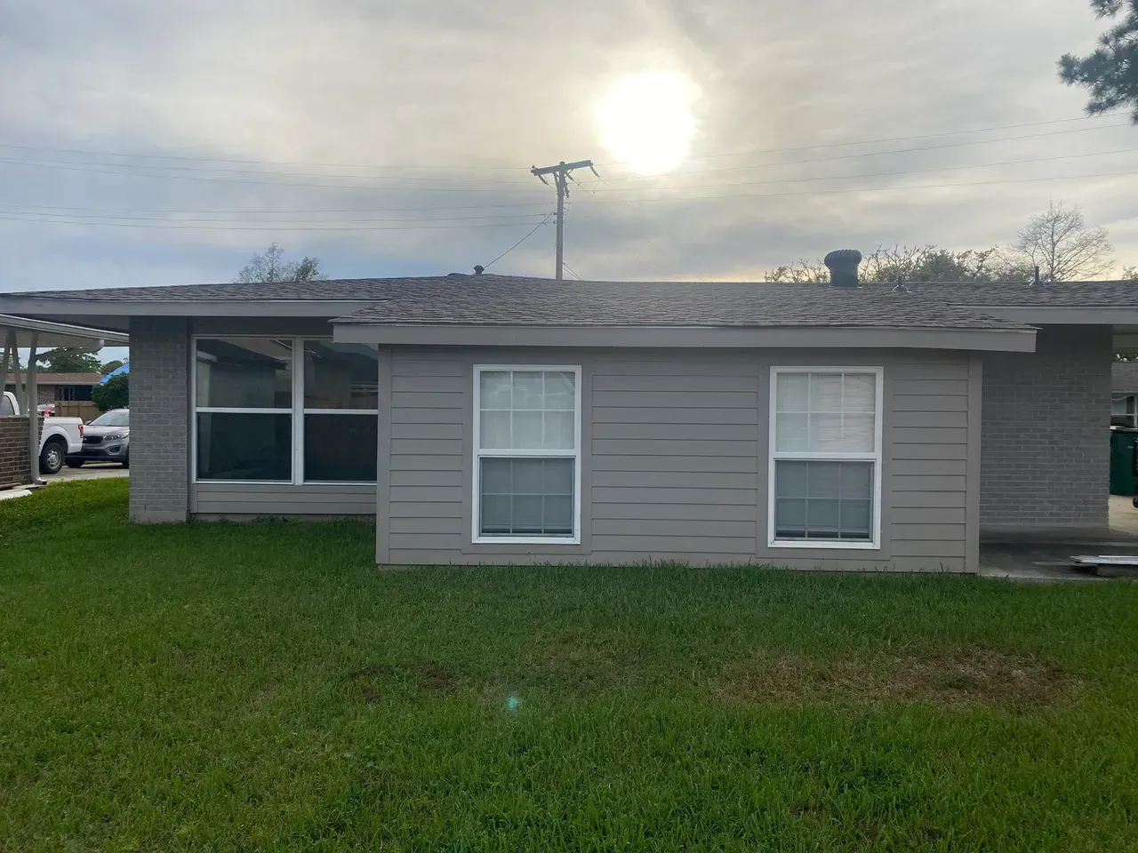 Gray house with two windows, large window, and green lawn under a cloudy sky.