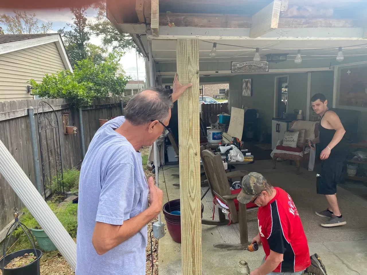 Three men working on a wooden porch support. One man looks on, another hammers, the third watches. Outdoors.