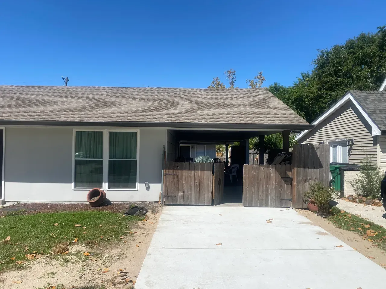 A low-slung house with a light gray exterior, wooden fence, and driveway under a clear blue sky.