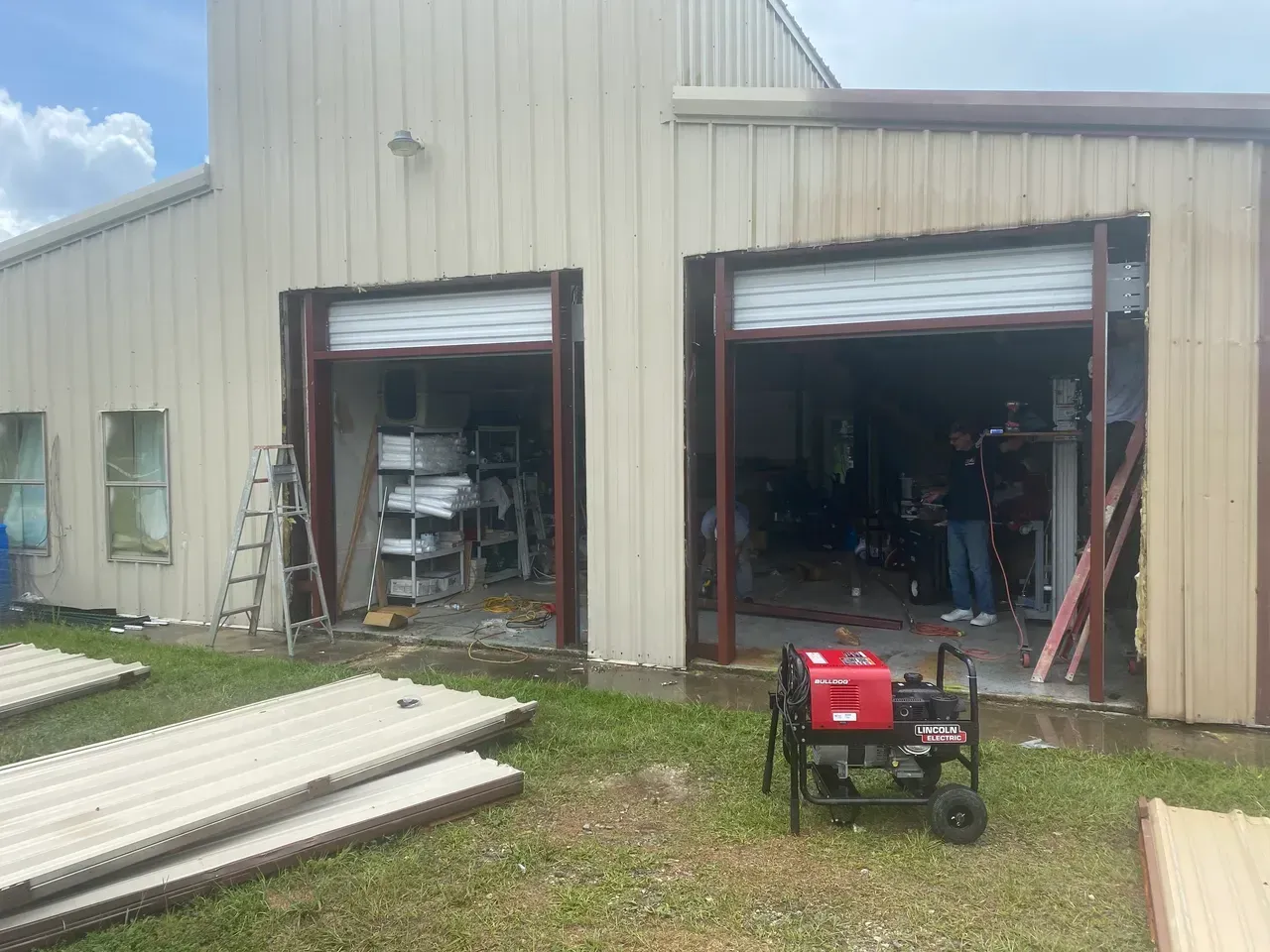 Exterior of a tan metal building with open garage doors; a generator is in front, and a person is visible inside.