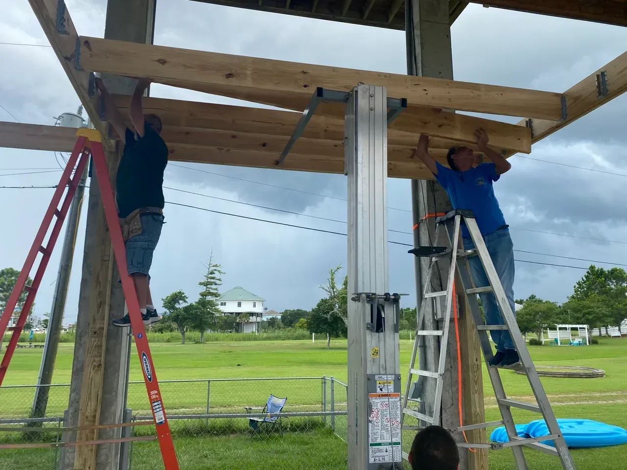 Two people on ladders installing wooden beams on a structure outdoors.