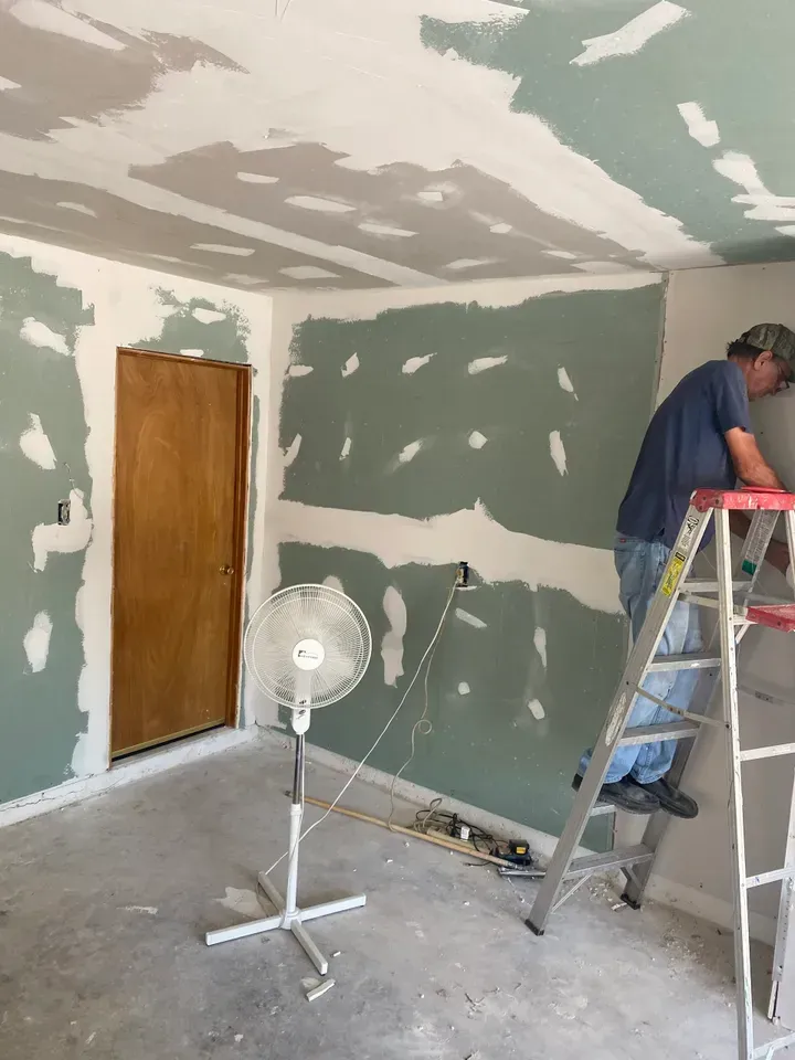A person on a ladder working on drywall in a room under construction; fan blowing.