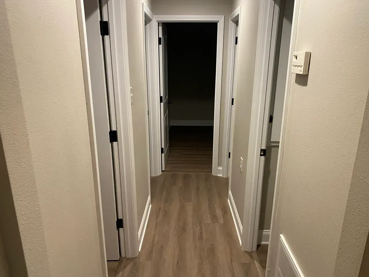 Hallway with doors on each side and light-colored flooring. The end door leads to a dark room.