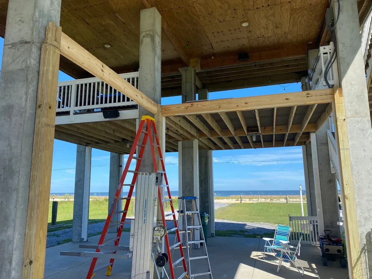 Coastal house under construction, elevated on concrete pillars with wooden framing. Ladders present.