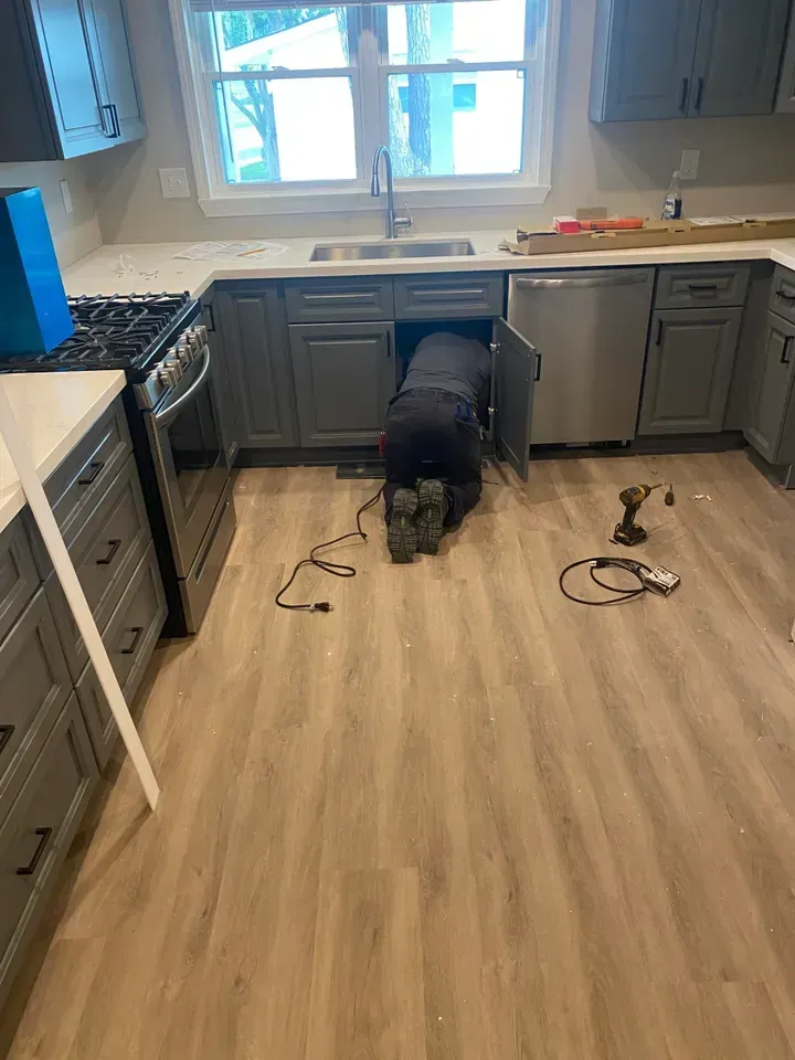 Person working under gray kitchen sink, tools on floor, light wood-look flooring.